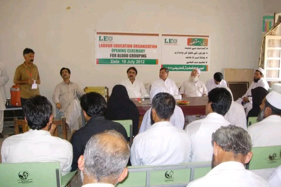 A full room of community members seated at the opening ceremony of a LEO blood grouping camp, with the organization's banner and a board of its aims on the wall