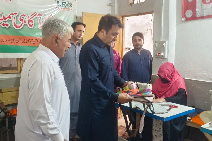 A woman working at a sewing machine in the skills centre at LEO's Mardan office, with LEO staff and visitors alongside
