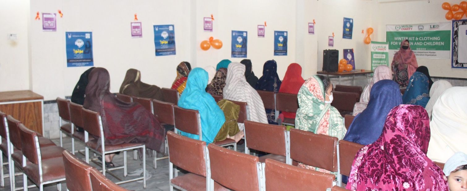 Women seated at a winter kit and clothing distribution session, seen from behind, as a speaker addresses the room beside the event banners