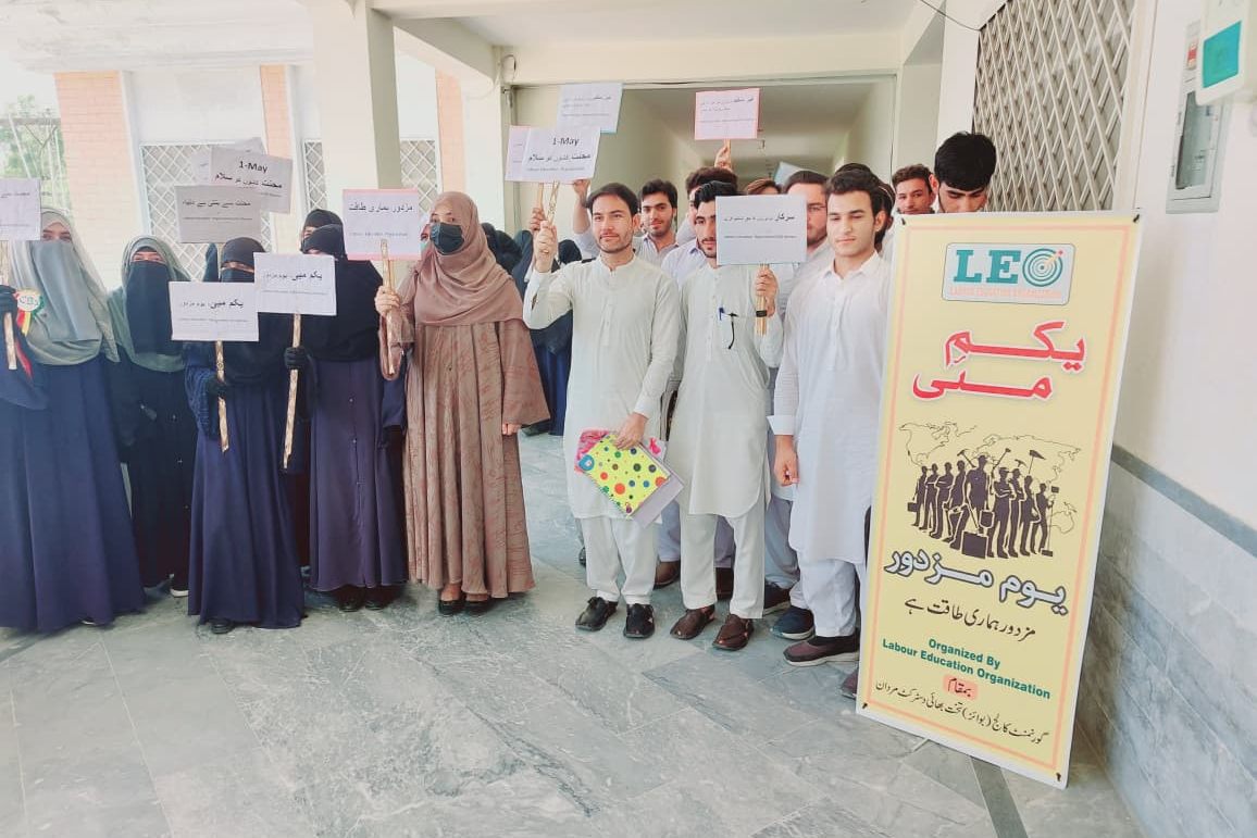 College students holding 1 May placards beside a LEO Workers' Day standee at Government College Takht Bhai